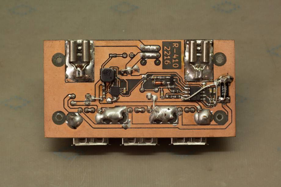 The bottom of the PCB, which has no solder mask. Visually dominating are a few large solder joints for the mechanical pins of the USB jacks and the 6.3 mm connectors (which are just regular crimp connectors originally made for cables). The main buck regulator IC is a tiny QFN near the centre of the PCB, surrounded by a slightly larger SMD inductor and a few capacitors. The top-mounted USB jacks can be seen protruding from behind the PCB at the bottom of the picture.
