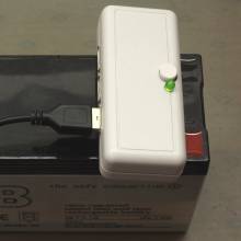The power bank in use: a small rectangular grey box is shown on top of a lead gel battery with a USB cable connected to one of the sockets on its left side. On its top right side, a small grey power switch and a green power indicator LED are poking through the case. The module is connected via the 6.3 mm contacts, which also holds it in place on top of the battery.